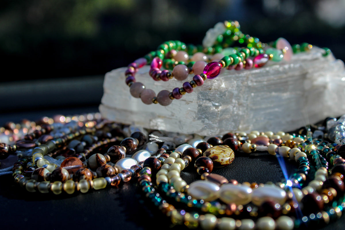 A close-up of multiple beaded bracelets and necklaces arranged on a dark surface, with a clear crystal slab holding a green and pink beaded bracelet in the background.